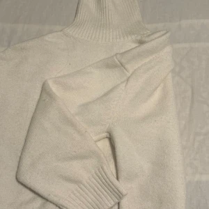 Vit polotröja i mjuk stickad ull - Klassisk vit polotröja i mjuk stickad ull med ribbade muddar och hög krage. Perfekt för lager-på-lager och ger en clean och stilren look. Passar dig som gillar minimalistisk stil och vill hålla dig varm under kyliga dagar.