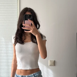 Vit ribbad croppad topp från H&M - Säljer en vit ribbad croppad topp från H&M med korta ärmar och rundad halsringning. Toppen är figurnära och har en enkel, stilren look som passar perfekt till jeans eller kjol. Materialet är mjukt och stretchigt för en bekväm känsla. Toppen har en liten fläck på.