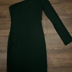 Mörkgrön ribbad one shoulder klänning - Snygg mörkgrön ribbad klänning med one shoulder-design och lång ärm. Tight passform som framhäver figuren och ger en modern look. Perfekt för dig som gillar stilrena plagg med en twist.