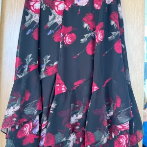 Svart blommig midi kjol  - Svart långkjol med stora röda och rosa blommor, volangdetaljer och slutar på vaden/något nedanför. Kjolen har en smal passform upptill och flödar sedan ut nertill i tunna våder lager på lager. Så snyggt till stövlar!
