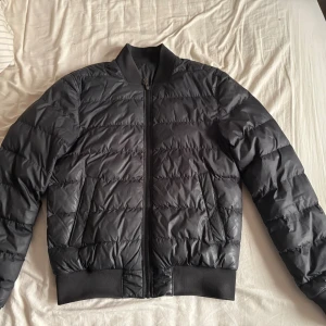 Svart j.Lindeberg jacka - Säljer en svart pufferjacka med ribbade muddar och ståkrage. Jackan har en klassisk quiltad look, två snedställda fickor och stängs med dragkedja framtill. Perfekt för kyliga dagar och ger en clean, modern vibe. Ordinarie pris 2999kr