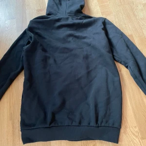 Svart hoodie från McKenzie med dragkedja - Svart hoodie från McKenzie med dragkedja framtill och snörning i huvan. Tröjan har långa ärmar och ribbade muddar vid ärmslut och nederkant. Perfekt för en chill och avslappnad stil.