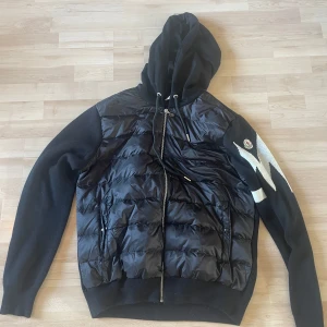 Svart pufferjacka med stickade ärmar Moncler - Snygg svart pufferjacka från Moncler med stickade ärmar och huva. Jackan har dragkedja framtill, två fickor och coolt vitt mönster på ena ärmen med Moncler-logga. Kroppen är quiltad och glansig medan ärmarna är i mjuk stickad kvalitet.