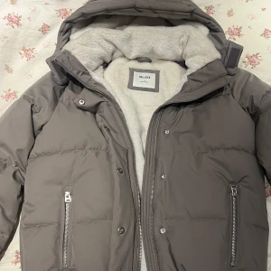 Grå pufferjacka från Hollister - Snygg grå pufferjacka från Hollister med mysigt ljust foder, stor huva och dragkedja framtill. Jackan har två fickor med dragkedja, ribbade muddar vid ärmsluten och justerbar huva med kardborre. Perfekt för kalla dagar och riktigt skön att bära. Normalpris från Hollister 1090kr/99€ (vilket är slutsåld just nu så passa på nu!!)