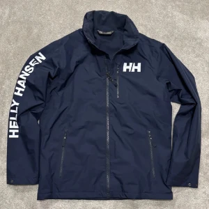 Marinblå vindjacka från Helly Hansen - Snygg bra vår/höst vindjacka från Helly Hansen  Jackan har dragkedja framtill, två fickor med dragkedja och hög krage + en luva som går att få ut. Perfekt för blåsiga dagar och sportig stil.