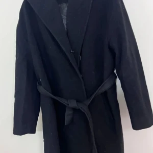 Svart kappa från Dixi Coat med bälte - Svart kappa från Dixi Coat med klassisk krage och knytbälte i midjan. Långärmad och helfodrad, perfekt för kyliga dagar. Stilren och enkel design som funkar till många outfits.