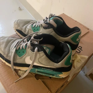 Nike Air Max sneakers grå och gröna - Nike Air Max sneakers i grått skinn med gröna och svarta detaljer. Klassisk vit swoosh på sidan, svart innersula och mesh vid plösen. Skorna har snörning och en platt sula med synlig Air Max-dämpning. Perfekta för dig som gillar retrostil och streetwear.