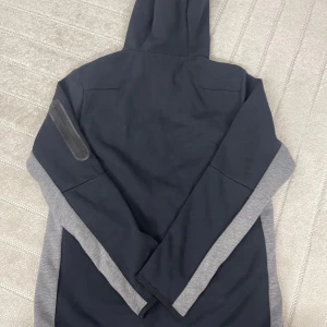 Nike hoodiejacka med dragkedja - Nike hoodiejacka i svart och grått med hel dragkedja och huva. Jackan har vita och grå ränder över bröstet, sidofickor och en liten ficka med dragkedja på ärmen. Tillverkad i bomull och polyester för en skön och sportig känsla.