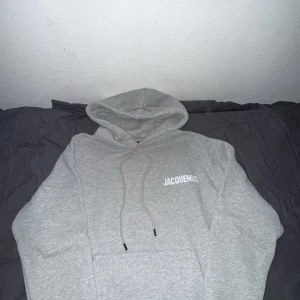 Grå hoodie från Jacquemus - Säljer en stilren grå hoodie från Jacquemus med vit logga på bröstet. Tröjan har huva med snörning och en klassisk känguruficka framtill. Perfekt för dig som gillar minimalistisk och trendig streetwear. Priset kan diskuteras;)