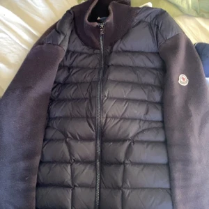 Svart dunjacka från Moncler - Säljer en svart dunjacka från Moncler med quiltad framsida och mjuka ärmar. Jackan har dragkedja framtill och en stilren design med Moncler-logga på ärmen. Perfekt för dig som vill ha en snygg och varm jacka med premiumkänsla.