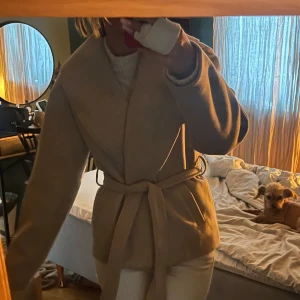 Beige kappa med bälte från ONLY XS - Snygg beige kappa från ONLY i kortare modell med bred krage och knytskärp i midjan. Jackan har en clean look och är perfekt för dig som gillar stilrena vibes. Tillverkad i mjukt material som känns lyxigt och passar till många outfits. Helt ny! Köptes för 699 kr.