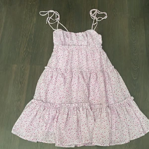 Blommig kortklänning från FB Sister - Supersöt kortklänning med volanger och smala axelband som knyts. Klänningen är ljuslila med små rosa blommor och har ett luftigt, flowy tyg med foder under. Perfekt för varma dagar och ger en romantisk vibe. Väldigt somrig och fin. Passar super fint till ballerina skor!! Eller tofflor😍😍