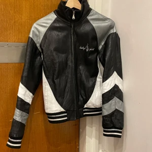 Baby Phat Skinnjacka - Snygg svart, vit och silver skinnjacka från Baby Phat med coola kontrastpaneler och broderad logga på bröstet. Jackan har dragkedja framtill, ribbade muddar och sportiga detaljer på ärmarna. Avtagbar fluff på insidan med dragkedja. Perfekt statement-plagg för dig som vill sticka ut. Super vintage och trendig!🫶