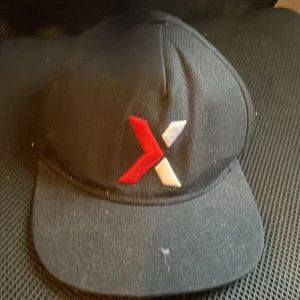 Svart keps med röd och vit logga - Svart keps med böjd skärm och justerbar snapback baktill. Framtill finns en broderad logga i rött och vitt som ger en cool kontrast. Kepsen är tillverkad i bomull och passar perfekt för en avslappnad streetstyle.
