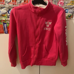 Rosa hoodie från Rosetti Riding SWE - Rosa hoodie från Rosetti Riding med dragkedja, broderade kronor och SWE på bröstet. Mjuk insida, två fickor fram och tryck på ena ärmen. Perfekt för dig som gillar ridning och vill sticka ut med en färgstark look.