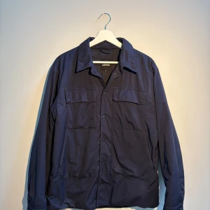 Field jacket från Aspesi - Stilren mörkblå overshirt från Aspesi med klassisk krage, tryckknappar och stora bröstfickor. Jackan har en enkel och clean look, perfekt för lager-på-lager. Tillverkad i lätt polyester som ger en smidig känsla och passar till många outfits.