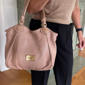 Beige handväska från Marc by Marc Jacobs - Säljer en snygg beige handväska från Marc by Marc Jacobs i äkta skinn. Väskan har guldiga metalldetaljer, dubbla handtag och en klassisk logoplatta framtill. Insidan är fodrad med tyg med diskret logomönster. Perfekt storlek för allt du behöver ha med dig.