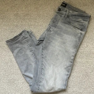 Grå  jeans från Jack & Jones - Snygga grå jeans från Jack & Jones i skinny fit med klassisk femficksdesign och diskret tvättad look. Jeansen har låg midja och är tillverkade i mjuk bomullsblandning med stretch för extra komfort. Perfekta för en avslappnad och trendig stil. 28/30