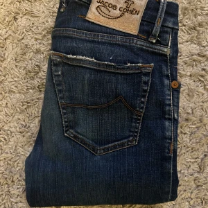 Mörkblå jeans från Jacob Cohen 622 - Snygga mörkblå jeans från Jacob Cohen med klassisk femficksdesign ex och läderpatch bak. Jeansen har raka ben och subtila slitningar vid fickorna för en cool look. Tillverkade i mjukt denimtyg med diskreta kontrastsömmar. Alla slitningar är design. Modell 622 ( slim fit )