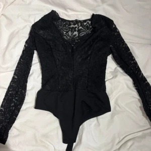 Svart spetsbody från NLY One XS - Säljer en svart body från NLY One i storlek XS. Bodyn har långa ärmar och är tillverkad i spets med blommigt mönster upptill och på ärmarna. Nederdelen är i slätare tyg. Perfekt för dig som vill ha en snygg och stilren topp med det lilla extra.