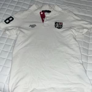 Vit pikétröja från Morris Company - Vit pikétröja från Morris Company med broderad logga och text på bröstet, svart siffra 8 på ärmen och röd knappslå. Klassisk krage och korta ärmar. Perfekt för en sportig och stilren look.