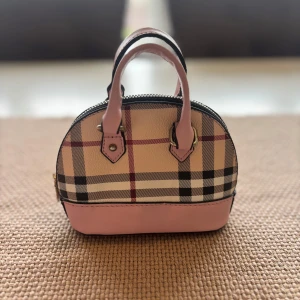 Rutig handväska med rosa detaljer - Söt liten handväska med klassiskt rutigt mönster och ljusrosa detaljer på handtag och botten. Väskan har rundad form, dragkedja upptill och är perfekt för det viktigaste. Materialet är skinnimitation och insidan är fodrad i svart tyg.