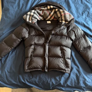 Burberry jacka/väst - Snygg svart pufferjacka från Burberry med avtagbara ärmar så den kan bäras som väst. Jackan har huva med klassiskt rutigt Burberry-foder, dragkedja och knappar framtill samt två fickor med dragkedja. Perfekt nu till hösten, kalla dagar och riktigt stilren. Helt oanvänd.