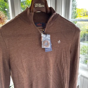 Brun merinoullströja från Morris - Snygg brun långärmad tröja från Morris i 100% merinoull. Tröjan har en stilren halv dragkedja vid halsen och en liten broderad logga på bröstet. Perfekt för dig som gillar klassisk och clean stil.