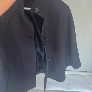 Svart poncho - Stilren svart cape-kappa med knappar framtill och diskreta fickor. Perfekt för dig som vill ha något annorlunda än en vanlig jacka. Stl 34/xs