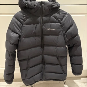 Svart dunjacka Peak Performance S - Säljer en svart dunjacka från Peak Performance i storlek S. Jackan har huva, dragkedja framtill och två sidofickor med dragkedja. Puffig modell som håller dig varm under kalla dagar. Klassisk logga på bröstet och ärmen.