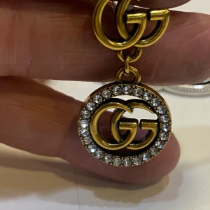 örhängen med kristaller -  örhängen i guldtonad metall med -loggor. Hängande cirkel med gnistrande klara kristaller runt loggan. Statement-accessoar som verkligen drar blickarna till sig. Perfekta för dig som älskar lyxiga detaljer och vill sticka ut.