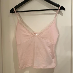 Ljusrosa linnetopp med spets Gina Tricot - Söt ljusrosa linnetopp från Gina Tricot med tunna axelband och v-ringning dekorerad med spets. Toppen är i mjuk bomull och har en feminin, enkel design som passar perfekt till jeans eller kjol.