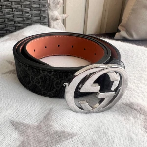 GG bälte - Snyggt svart bälte från GG med klassiskt GG-mönster och stort silvrigt GG-spänne. Bältet är gjort i skinn med insida i brun nyans och har flera hål för justering. Perfekt accessoar för att lyfta din outfit med en lyxig touch.