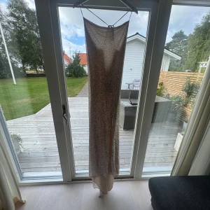 Glittrig långklänning med slits - Säljer en superfin långklänning i champagnefärgad ton med glittriga paljetter över hela klänningen. Klänningen har smala axelband, öppen rygg med knyt och en snygg slits framtill. Perfekt för dig som vill sticka ut och glänsa på festen! Köpt för 2500 prislapp kvar