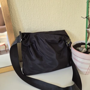 Svart axelväska från Åhlens - En stilren svart axelväska i polyester med justerbar rem och dragkedja upptill. Väskan är rymlig nog för en laptop och har flera innerfack för småprylar. Perfekt för dig som vill ha en snygg väska till skolan! 