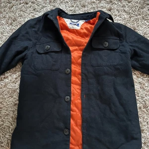 Svart overshirt från Jack & Jones - Stilren svart overshirt från Jack & Jones med orange quiltat foder. Jackan har två bröstfickor med lock och knappar framtill. Perfekt för lager-på-lager och har en klassisk krage och lång ärm.
