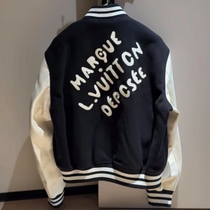 Louis Vuitton svart och vit bomberjacka - Snygg svart och vit bomberjacka från Louis Vuitton med broderade detaljer och pärlor runt LV-loggan. Jackan har vita ärmar i skinn, svarta muddar med vita ränder och tryckknappar framtill. Baksidan har stor broderad text. Perfekt statement-plagg för dig som vill sticka ut.