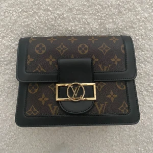 Brun axelväska  - Snygg axelväska, klassiskt monogrammönster med svarta skinnkanter och guldfärgade detaljer. Väskan har både kedja och läderrem som kan bytas ut, samt ett rymligt inre med flera fack. Perfekt för dig som vill ha en ikonisk och trendig accessoar. Osäker på äkthet. 