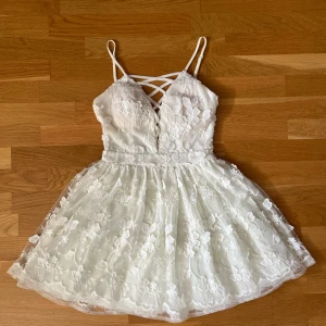 Vit blommig spetsklänning med snörning - Supersöt vit kortklänning med tunna axelband och snörning framtill. Klänningen har ett lager av transparent spets med broderade blommor och en v-ringad urringning. Kjolen är voluminös och bak finns en dragkedja för enkel påklädning. Köptes i Kanada och använd en gång till studenten!
