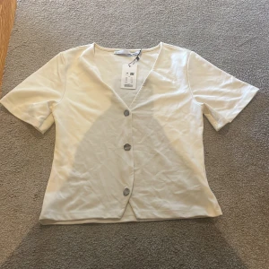 Vit kortärmad blus med knappar XS - Snygg vit blus från Lager 157 i storlek XS. Modellen har v-ringning, tre dekorativa knappar framtill och korta ärmar. Tillverkad i mjukt bomullstyg, perfekt för en clean och stilren look.