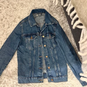 Klassisk blå jeansjacka - Säljer en klassisk blå jeansjacka med knappar framtill och två bröstfickor med lock. Jackan har en rak passform och långa ärmar, perfekt för dig som gillar en tidlös och avslappnad stil.