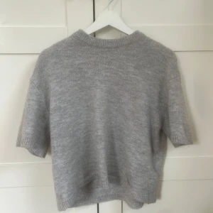 Grå stickad kortärmad tröja - Mysig grå stickad tröja med korta ärmar och rund hals. Perfekt för lager-på-lager och passar till både jeans och kjol. Enkel och stilren design med ribbade kanter vid ärmslut och nederkant.