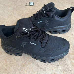 Svarta On Cloudrock Waterproof sneakers - Säljer ett par svarta On Cloudrock Waterproof sneakers med robust sula och vattentätt material. Skorna har snörning, diskret logga på sidan och är designade för att klara tuffare terräng. Perfekta för dig som vill ha både stil och funktion i ett par skor.