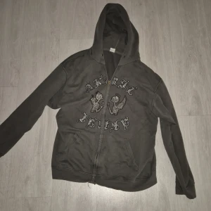 Mörkgrå hoodie med ängeltryck - Säljer en mörkgrå rhinestone hoodie med dragkedja och huva. Framsidan har ett stort tryck med två änglar och cool text i glittriga detaljer. Hoodien har känguruficka och långa ärmar, perfekt för en chill och avslappnad stil.
