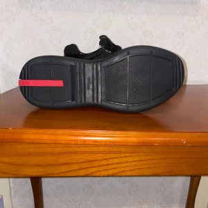 Svarta sneakers från Prada - Snygga svarta sneakers från Prada med en mix av glansig skinn och mesh. Skorna har svarta snören, rund tå och en robust svart sula med röd Prada-detalj på hälen. Perfekt för dig som gillar stilrena och exklusiva sneakers med en sportig touch.