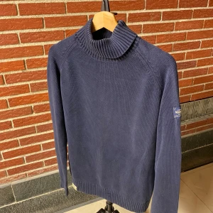 Mörkblå polotröja från Morris Division - Snygg mörkblå polotröja från Morris Division med hög ribbad krage och diskret patch på ärmen. Tröjan är stickad i ett mjukt material och har långa ärmar. Perfekt för dig som gillar stilren och klassisk look.