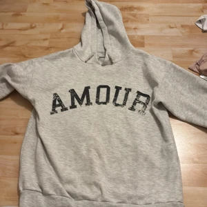 Grå hoodie med tryck från Gina Tricot - Grå hoodie från Gina Tricot med svart texten 'AMOUR' framtill i sliten stil. Tröjan har huva och ribbade muddar. Perfekt för chill dagar och enkel att matcha med jeans eller leggings.