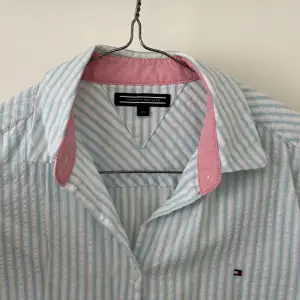 Superfin och nästan helt oanvänd Tommy Hilfiger skjorta i storlek 176. I linnetygsliknande material och väldigt tunn och svalkande till sommaren. Jättesöt ljusblå färg med rosa detaljer och vita ränder. 