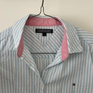 Tommy Hilfiger skjorta - Superfin och nästan helt oanvänd Tommy Hilfiger skjorta i storlek 176. I linnetygsliknande material och väldigt tunn och svalkande till sommaren. Jättesöt ljusblå färg med rosa detaljer och vita ränder. 