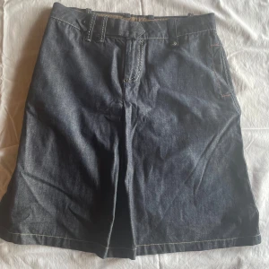 Mörkblå jeanskjol - En mörkblå jeanskjol med bälteshällor och framfickor. Kjolen har en knapp och dragkedja framtill samt kontrastsömmar.